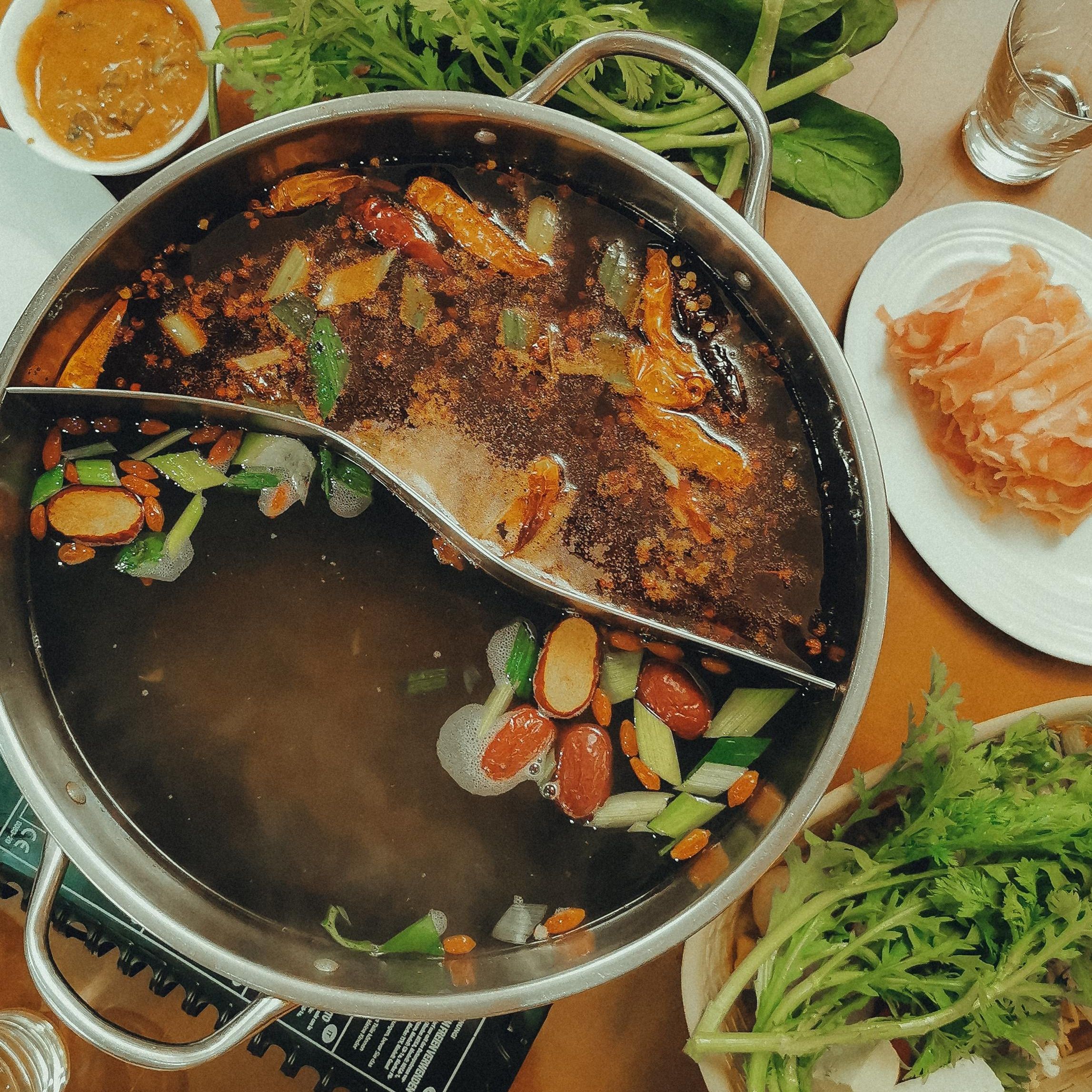 En hotpot med två sektioner med grönsaker, kryddor och kött. Det står tallrikar och skålar med kött, såser och grönsaker kring grytan.